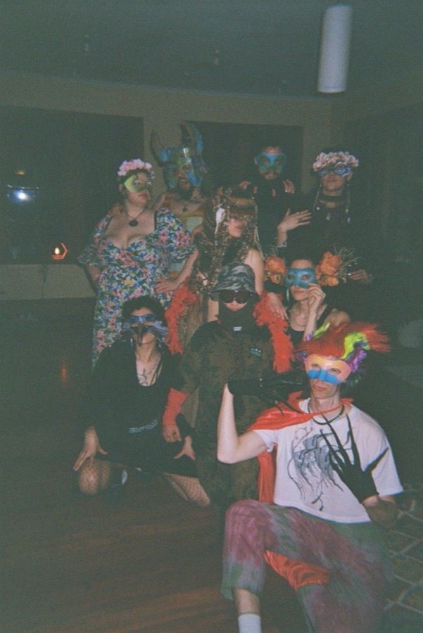 A big group of masked guests in feathers, flower crowns and capes posing together in a dim room