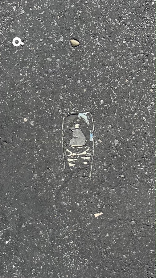 An outline shaped exactly like an old Nokia phone worn flat into dark grey road asphalt, scraps of grey screen and cream keypad still inside it