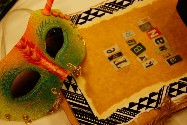 A glittery green and orange horned mask on kraft paper edged with black and white triangle patterns and cut-out magazine letters