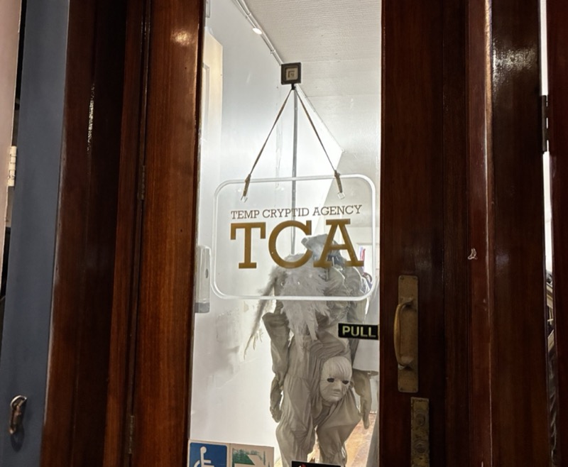 Wooden door with a hanging clear sign reading “TEMP CRYPTID AGENCY / TCA”, a pale masked costume figure behind the glass