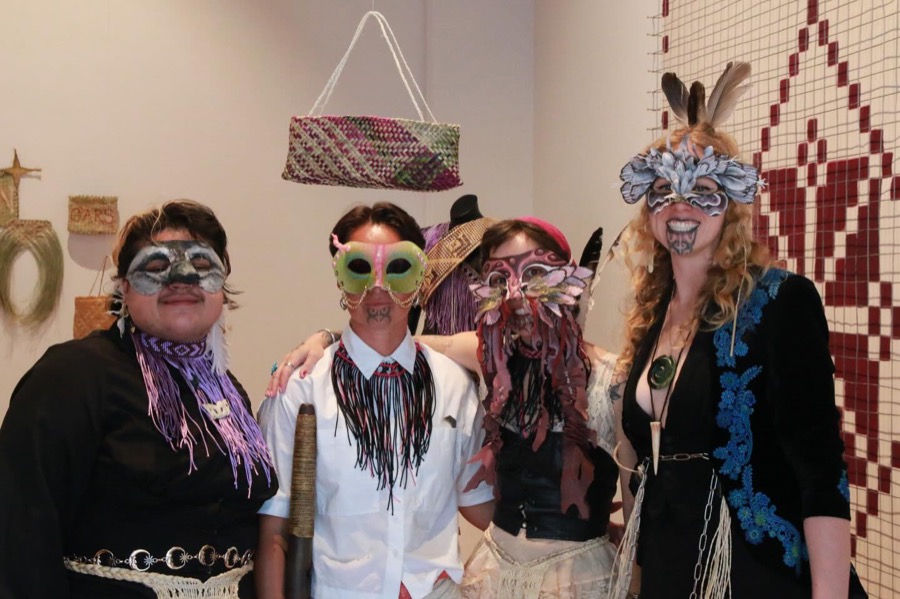Four people in handmade masks pose under a hanging woven kete in a gallery, one with feathers, one with pink leather tendrils