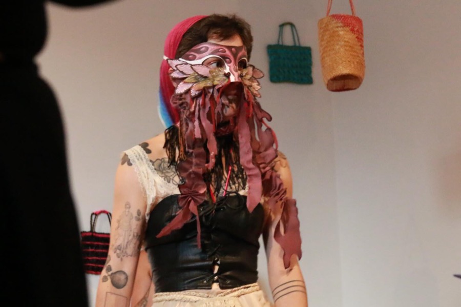 A person in a pink leather mask with long dangling tendrils glances sideways in a gallery hung with woven bags