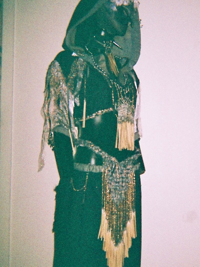 A hooded costume on a mannequin, dark fabric with silver sleeves, gold beaded chains and pale fibre tassels, shot on grainy film
