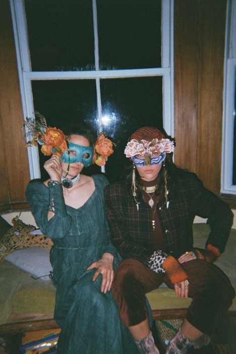 Seraphina and Skankenstein on a window seat at night, one in a teal mask with orange flowers, one in a flower-crowned blue mask and plaid jacket