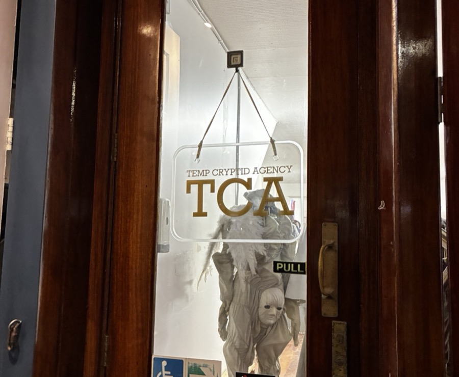 Wooden door with a hanging clear sign reading "TEMP CRYPTID AGENCY / TCA", a pale masked costume figure behind the glass